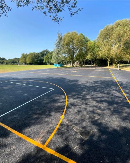 A sunlit basketball court with visible markings and trees in the background.